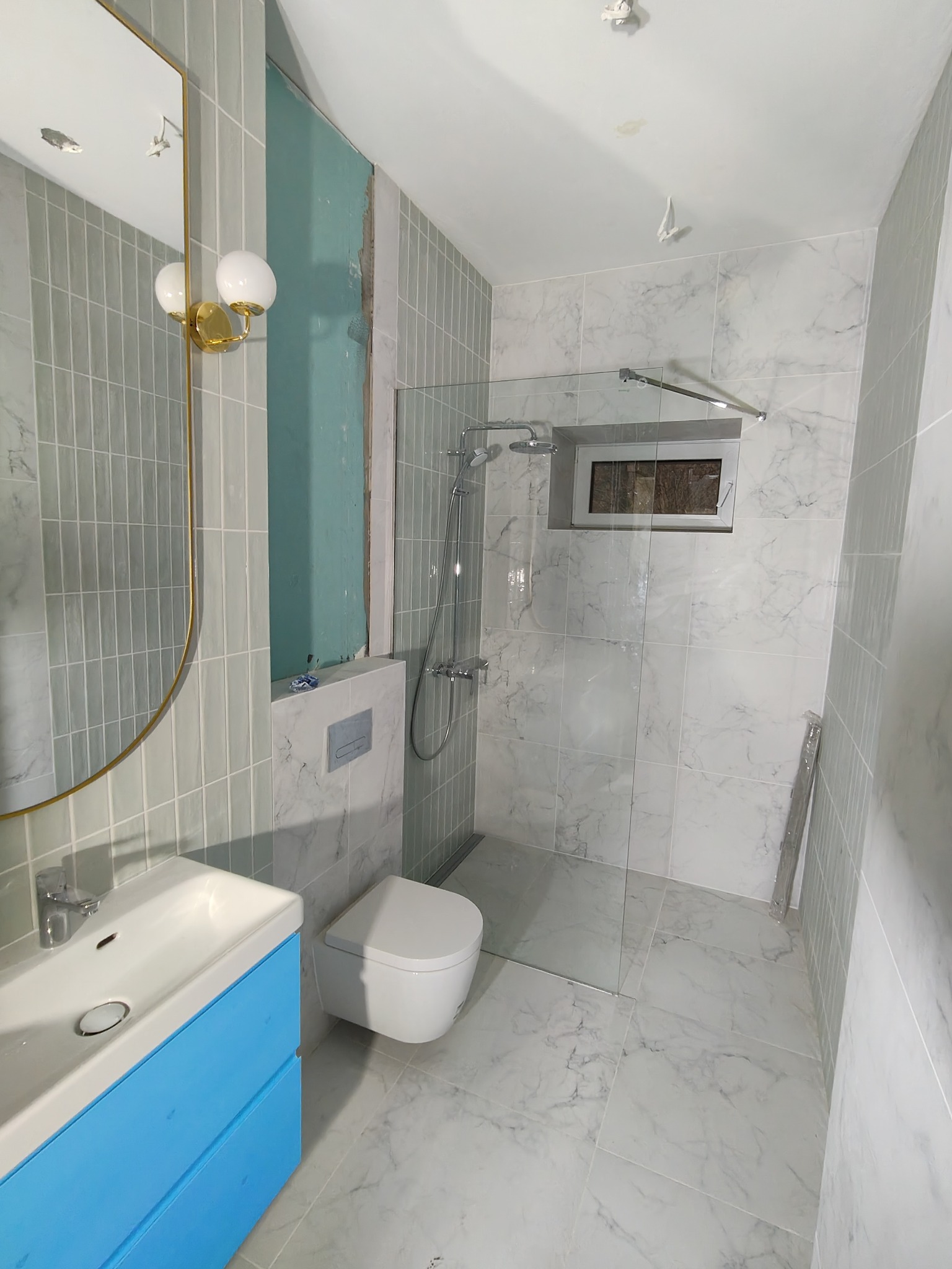 Jasna łazienka z kabiną prysznicową typu walk-in, umywalką z niebieską szafką i toaletą. Ściany wyłożone płytkami imitującymi marmur. Złote detale lustra i oświetlenia.