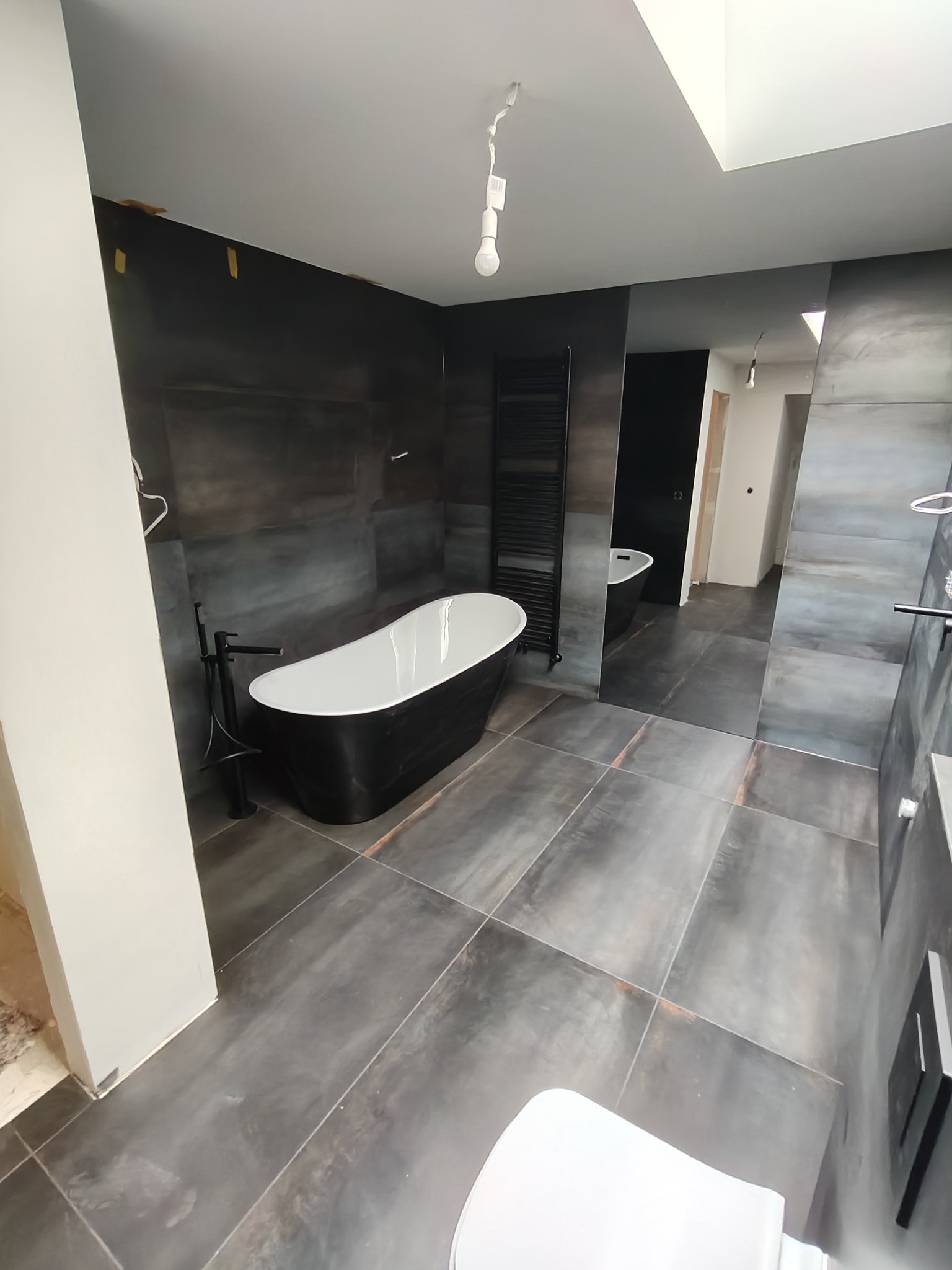 Nowoczesna łazienka z wolnostojącą wanną w czarno-białej kolorystyce, ciemnymi płytkami imitującymi beton i minimalistycznym grzejnikiem drabinkowym. Widok na lustro.