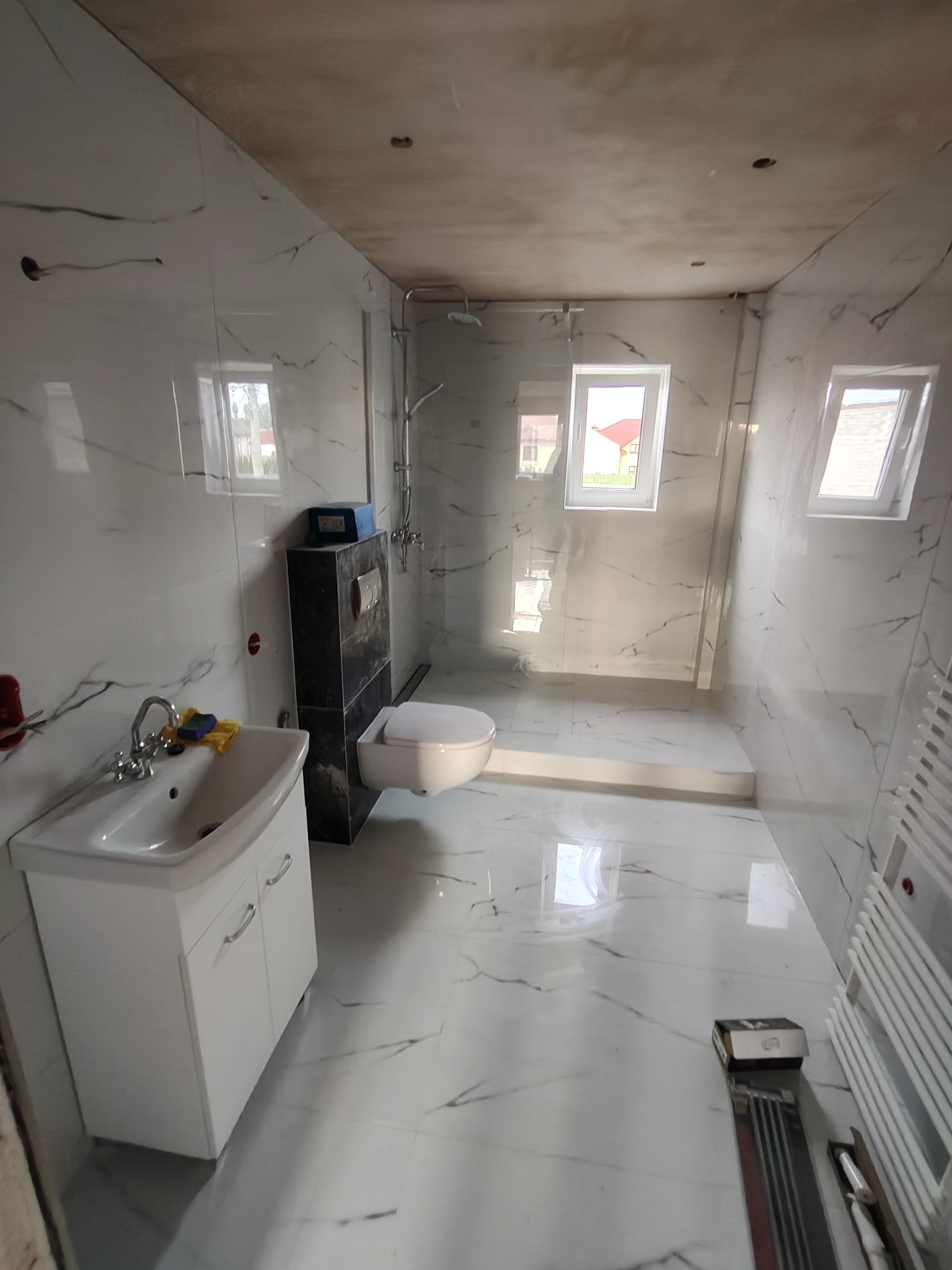 Jasna łazienka z marmurowymi płytkami, białą armaturą i kabiną prysznicową typu walk-in. Widoczne okna i niedokończony sufit.