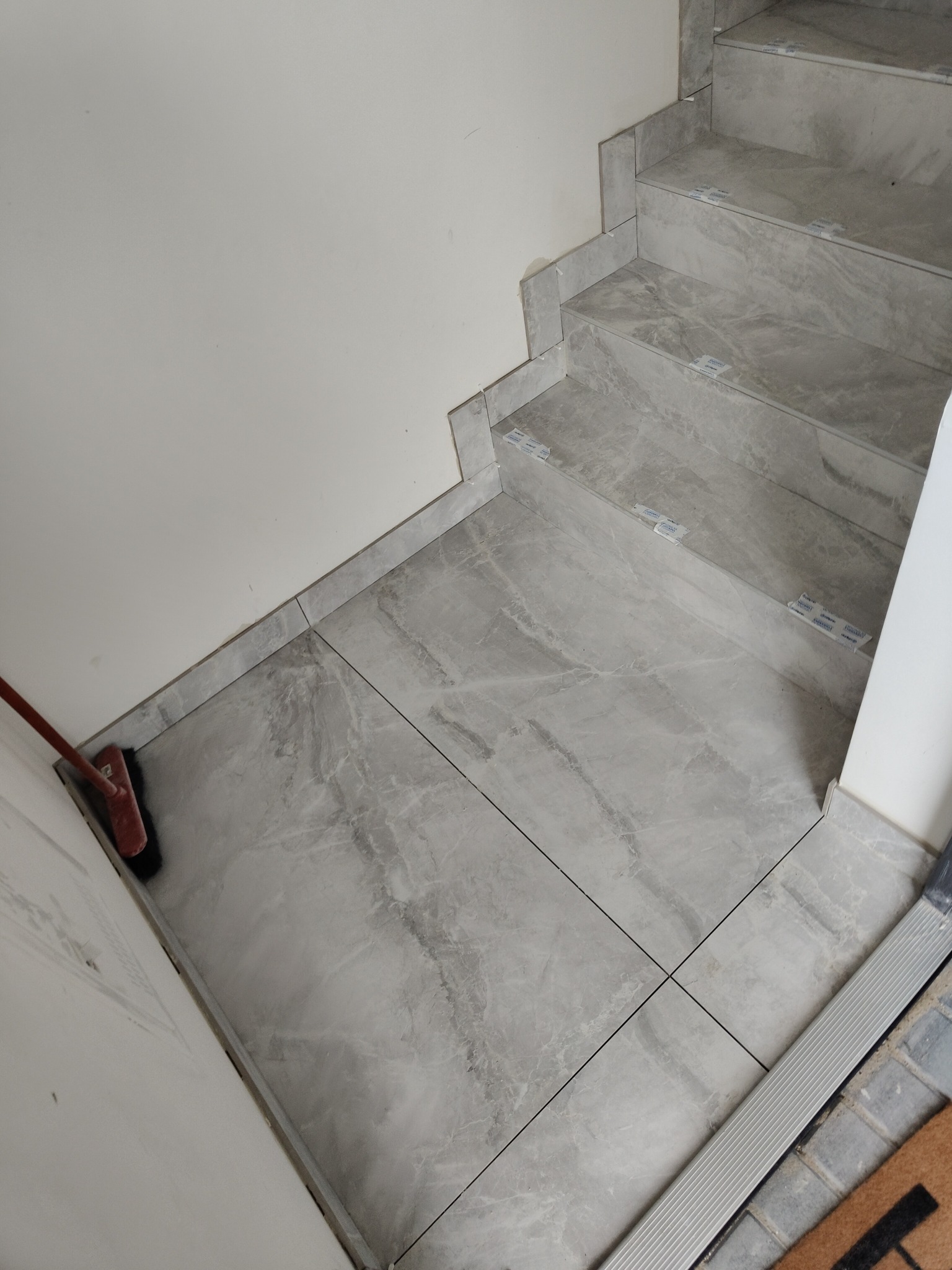 Wykończone schody i podłoga z szarych płytek imitujących marmur, widoczne krzyżyki dystansowe, ściana pomalowana na biało, w rogu stoi miotła.