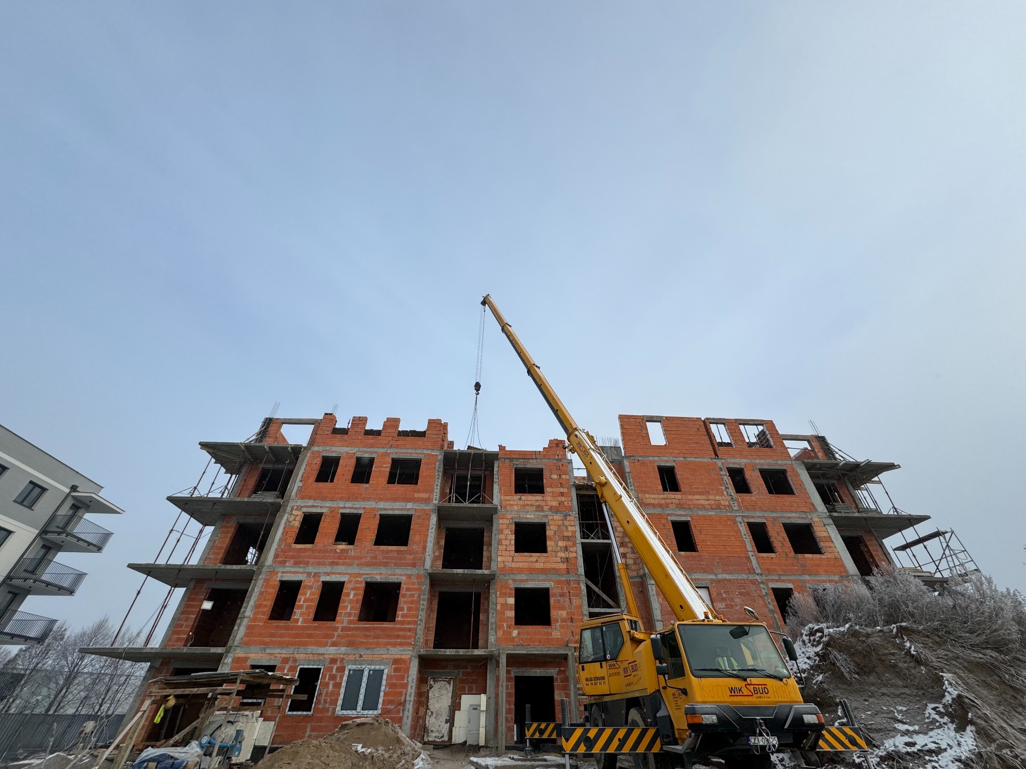 Budowa bloku z czerwonej cegły w trakcie wznoszenia, z widocznym żółtym dźwigiem. Ujęcie z dołu, pochmurne niebo w tle. Widoczny fragment innego budynku.