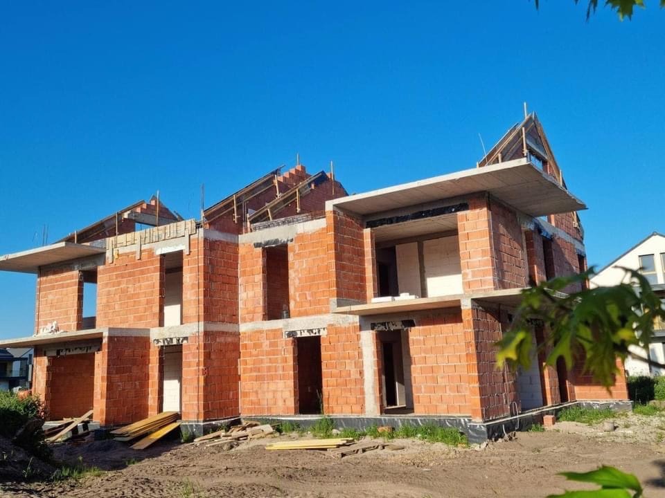 Wznoszenie dwukondygnacyjnego domu z czerwonej cegły, widoczne otwory okienne i zbrojenia stropów, błękitne niebo w tle. Etap surowy otwarty.