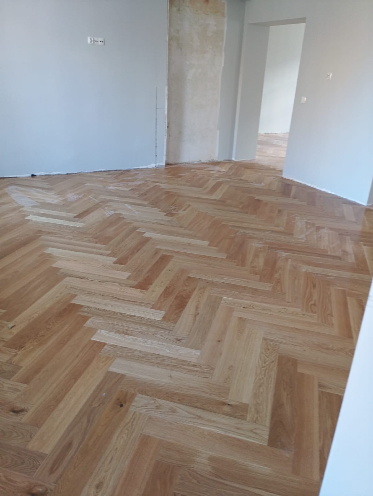 Ukończona podłoga z parkietu ułożonego w jodełkę w jasnym pomieszczeniu z szarymi ścianami. Widoczny fragment ściany w stanie surowym. W tle otwarte drzwi.