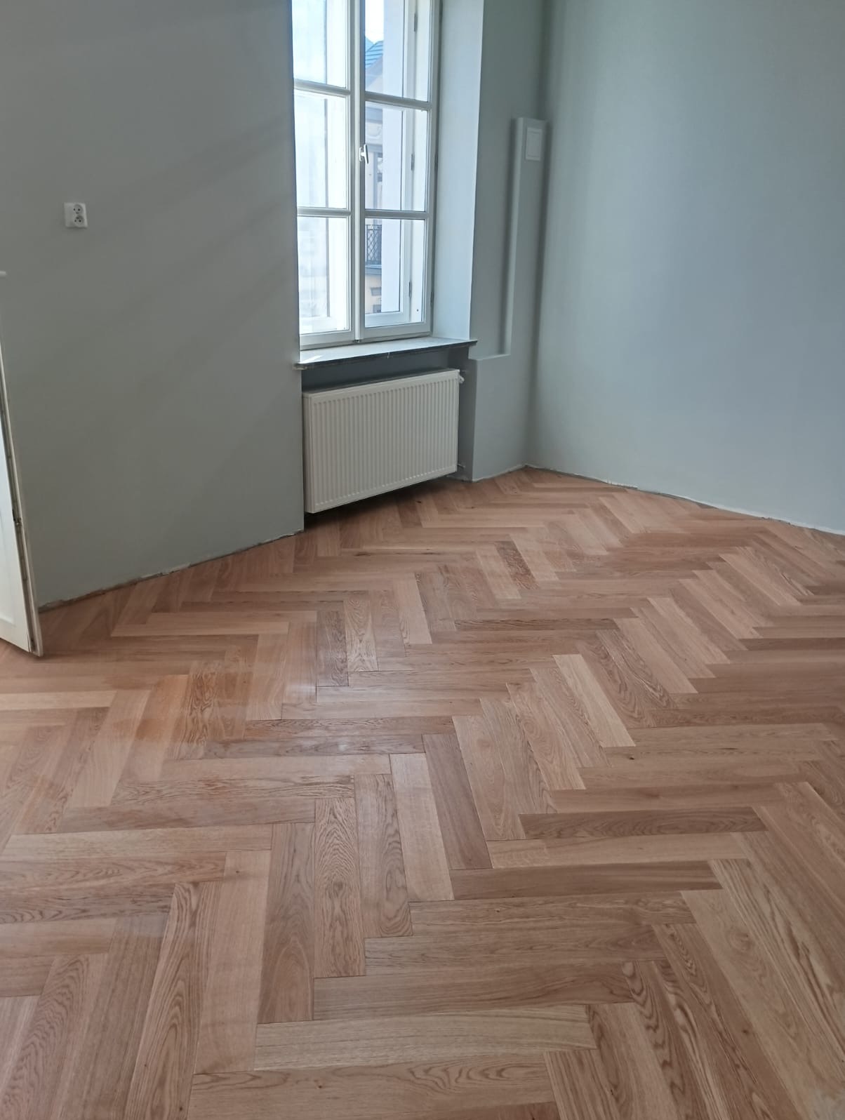 Ukończona podłoga z parkietu ułożonego w jodełkę w jasnym, odnowionym pokoju z oknem i grzejnikiem. Świeżo pomalowane ściany w odcieniach szarości.