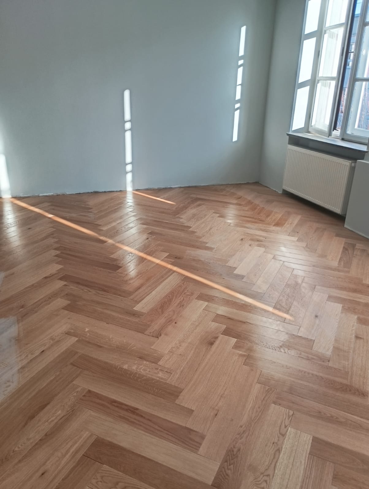 Jasna, drewniana podłoga ułożona w jodełkę w pustym pokoju z grzejnikiem pod oknem. Na ścianie widoczne smugi światła słonecznego.