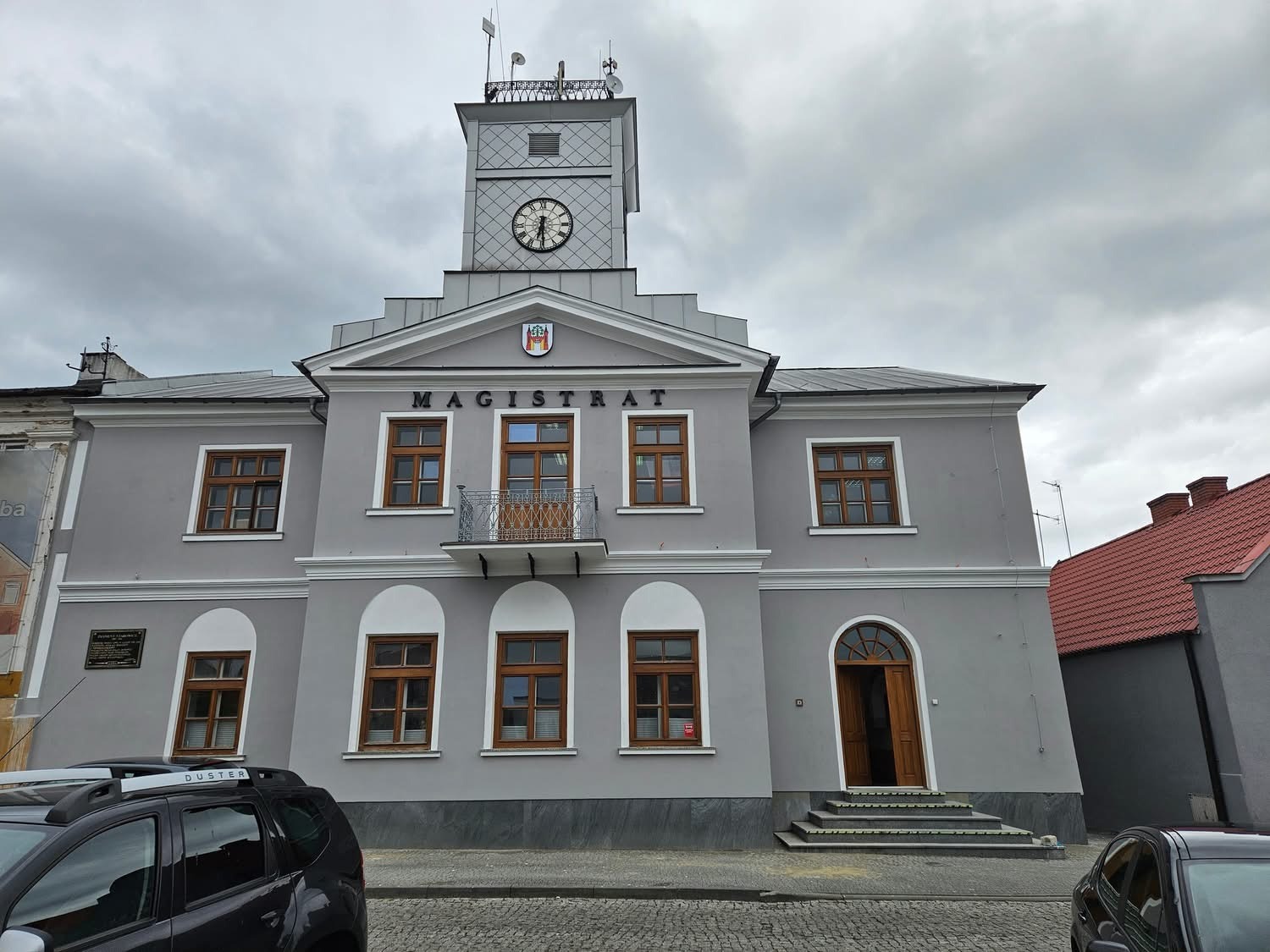Szary budynek magistratu z wieżą zegarową, balkonem i herbem. Widoczne okna z brązowymi ramami, fragment samochodu i kostka brukowa. Pochmurne niebo w tle.