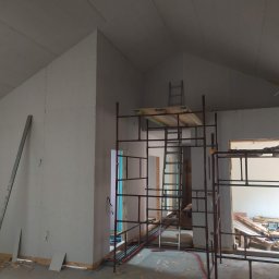 Vasyl Lutchyn - Montaż płyt karton-gips na poddaszu. Widoczne rusztowania, narzędzia i częściowo widoczna osoba. Ściany i sufit pokryte płytami, gotowe do dalszych prac wykończeniowych.