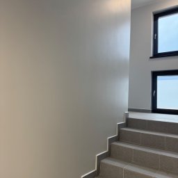 Vasyl Lutchyn - Klatka schodowa z szarymi płytkami i świeżo pomalowaną ścianą w odcieniach szarości. Dwa okna z czarnymi ramami w tle. Minimalistyczny design.