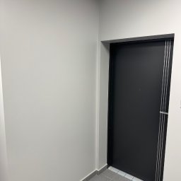 Vasyl Lutchyn - Nowoczesne, czarne drzwi wejściowe z metalowymi akcentami w jasnym korytarzu. Minimalistyczny design, szare płytki podłogowe, świeżo pomalowane ściany.