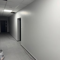 Vasyl Lutchyn - Szary korytarz po malowaniu z widocznymi drzwiami, szarą podłogą z płytek i elementami instalacji pod sufitem. Świeżo pomalowane ściany w neutralnym odcieniu szarości.