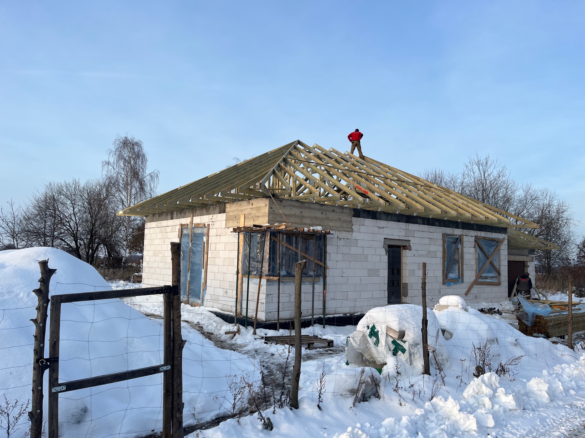 Wznoszenie domu z białego bloczka: widoczna więźba dachowa, pracownik na dachu w czerwonej kurtce. Zimowy krajobraz z zaspami śniegu.