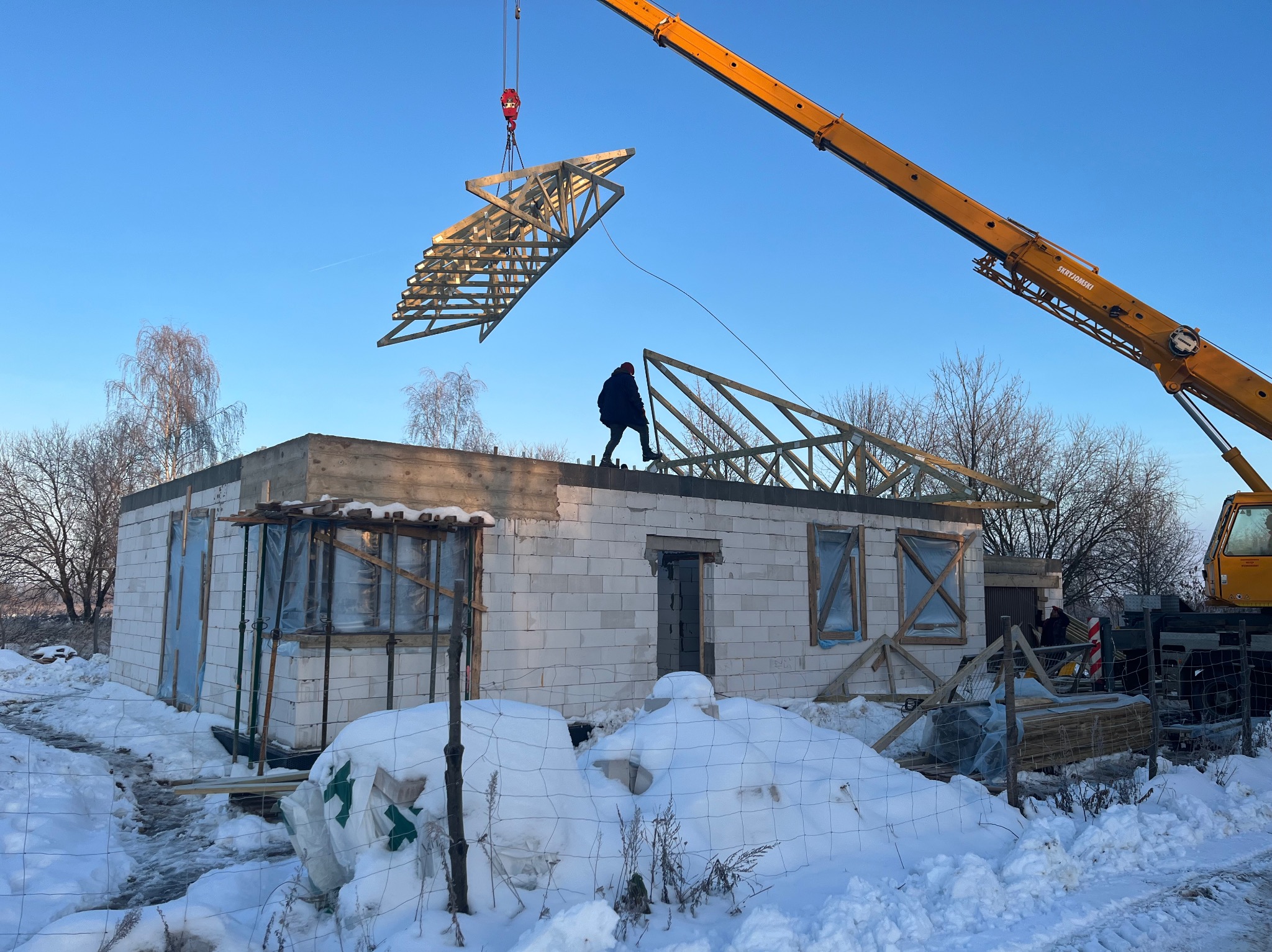 Montaż więźby dachowej na domu z białych bloczków, z użyciem dźwigu w zimowej scenerii. Widoczny pracownik na dachu i elementy konstrukcyjne.