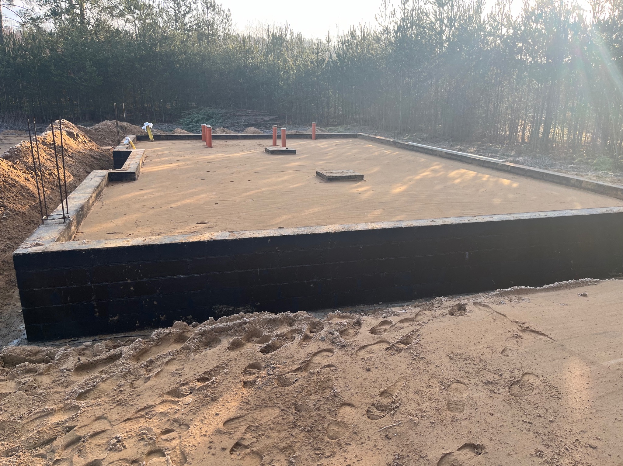 Zalane fundamenty domu jednorodzinnego, widoczne zbrojenia i pionowe rury instalacyjne. W tle las iglasty w porannym słońcu.