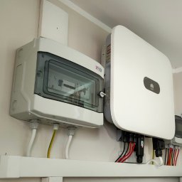 PSK Sp. z o.o. - Biała skrzynka rozdzielcza ETI z przezroczystą pokrywą i falownik Huawei zamontowane na ścianie. Widoczne przewody elektryczne.