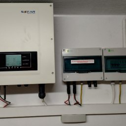 PSK Sp. z o.o. - Biały inwerter solarny Sofar Solar z wyświetlaczem i podłączonymi kablami, obok dwie skrzynki elektryczne na białej ścianie. Widoczne rury instalacyjne.