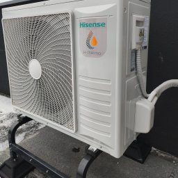 PSK Sp. z o.o. - Biała jednostka zewnętrzna klimatyzacji Hisense Hi-Therma na czarnym stelażu, z widoczną kratką wentylatora i rurą odprowadzającą, na tle szarej ściany i fragmentu śniegu.
