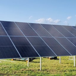 PSK Sp. z o.o. - Panel słoneczny na metalowej konstrukcji, zainstalowany na trawniku przed domem jednorodzinnym, na tle błękitnego nieba. Widoczne drzewa i ogrodzenie.