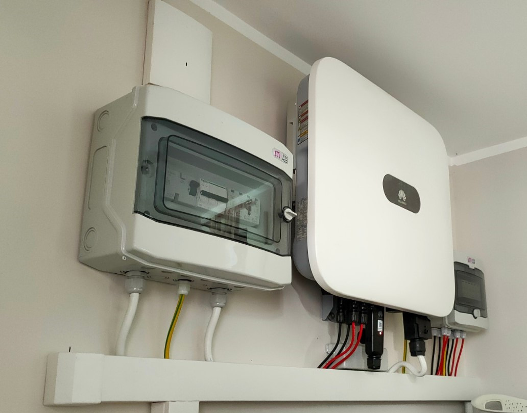 Biała skrzynka rozdzielcza ETI z przezroczystą pokrywą i falownik Huawei zamontowane na ścianie. Widoczne przewody elektryczne.