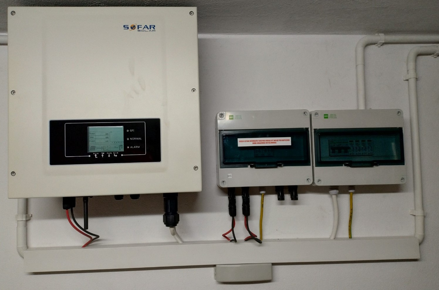 Biały inwerter solarny Sofar Solar z wyświetlaczem i podłączonymi kablami, obok dwie skrzynki elektryczne na białej ścianie. Widoczne rury instalacyjne.