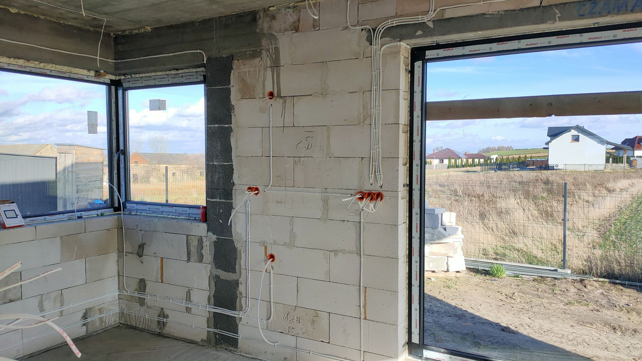 Wnętrze w stanie surowym z widoczną instalacją elektryczną: białe przewody prowadzone po ścianach z betonu komórkowego, pomarańczowe puszki pod gniazdka i włączniki.