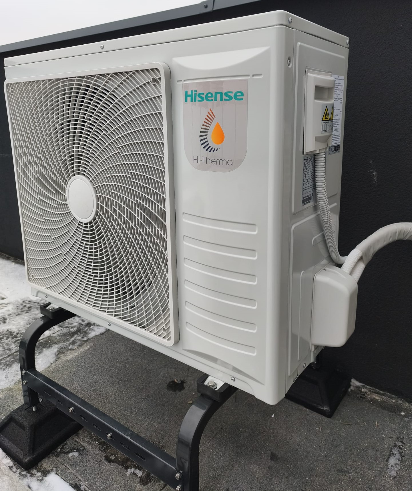 Biała jednostka zewnętrzna klimatyzacji Hisense Hi-Therma na czarnym stelażu, z widoczną kratką wentylatora i rurą odprowadzającą, na tle szarej ściany i fragmentu śniegu.