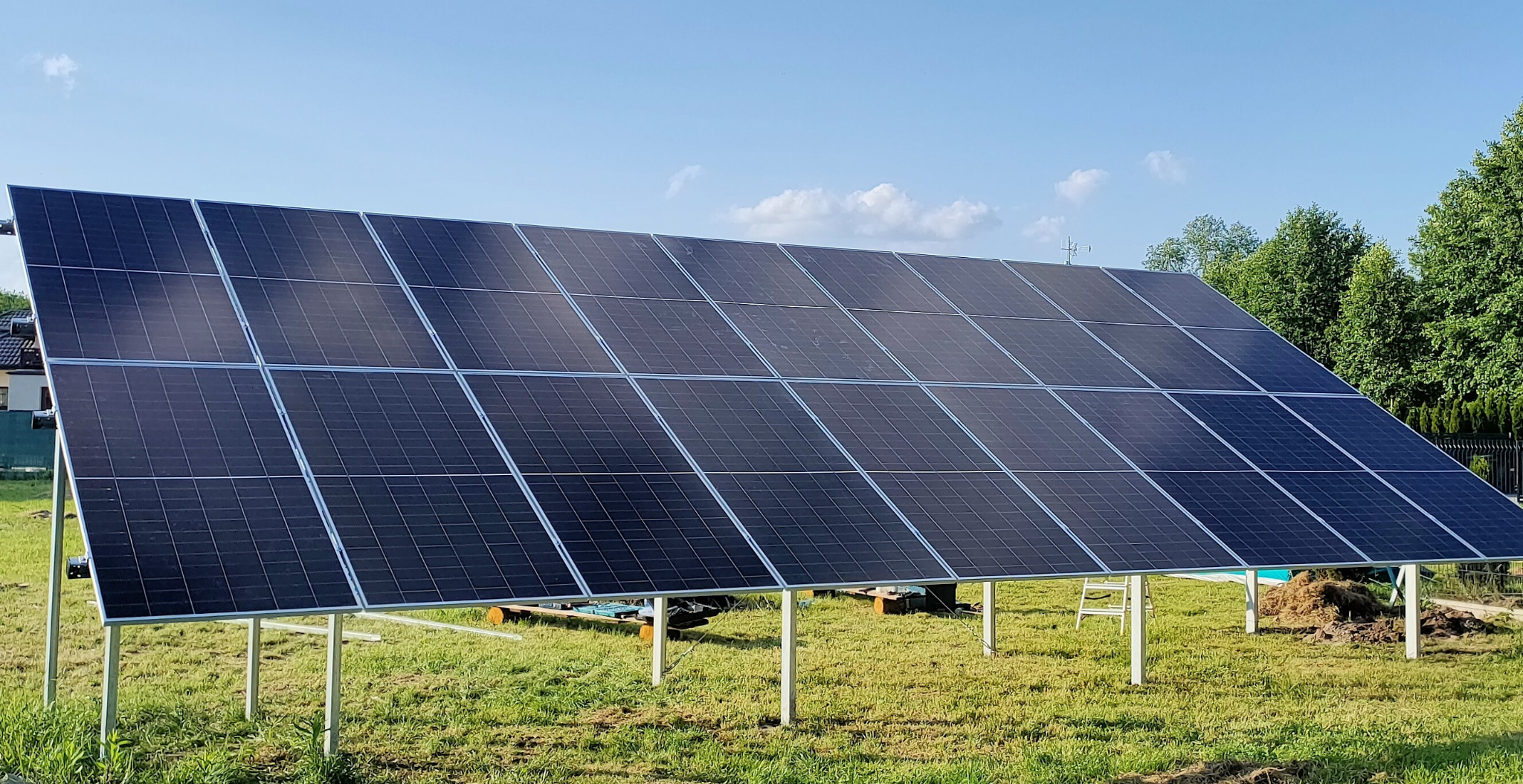 Panel słoneczny na metalowej konstrukcji, zainstalowany na trawniku przed domem jednorodzinnym, na tle błękitnego nieba. Widoczne drzewa i ogrodzenie.