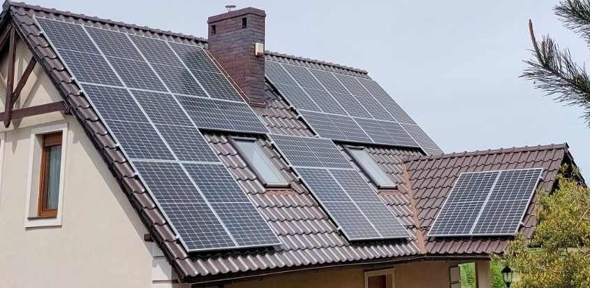 Panele słoneczne na dachu domu z brązową dachówką, widoczne okna dachowe i komin. Instalacja fotowoltaiczna na skośnym dachu.