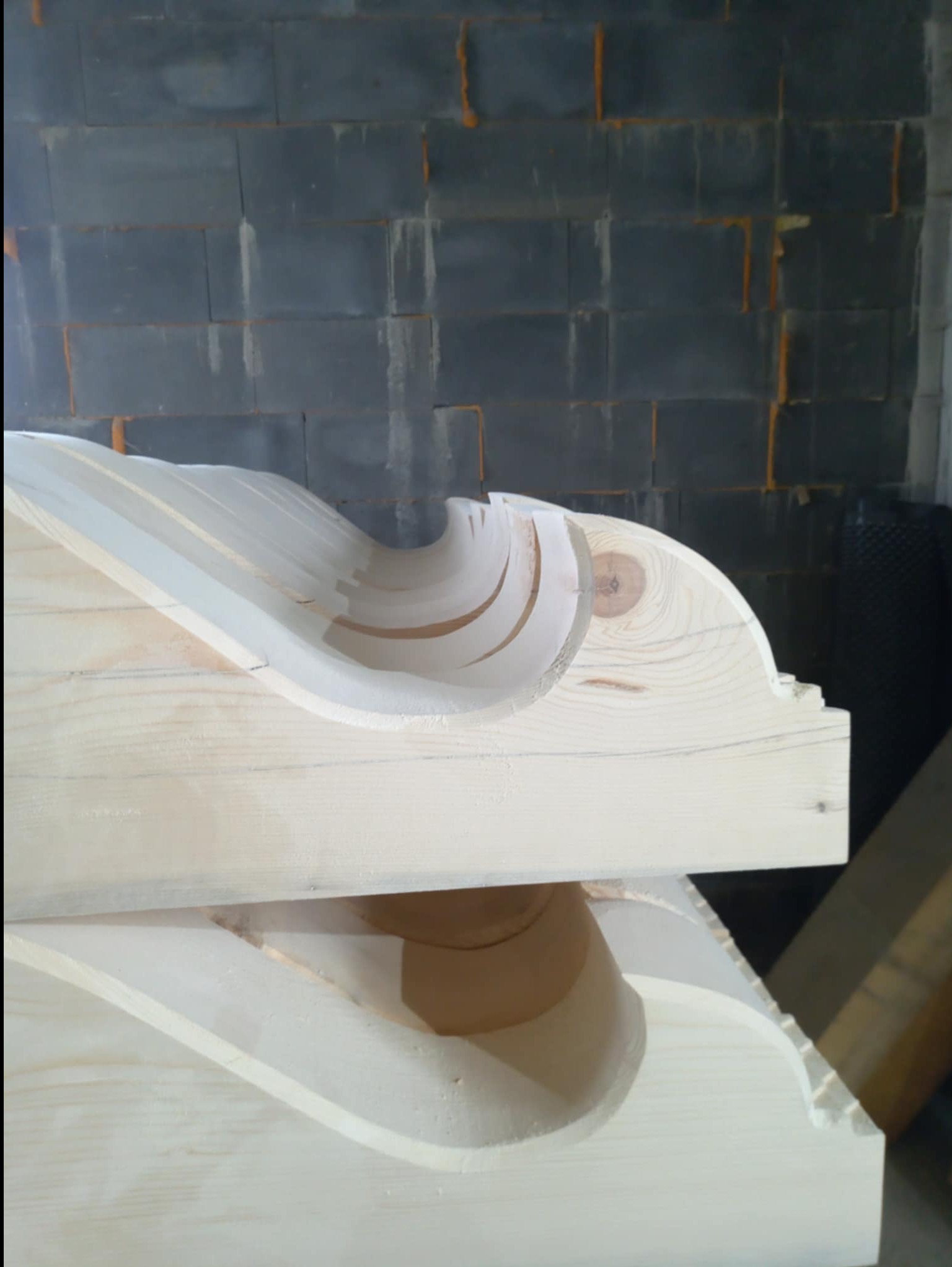 Drewniany, rzeźbiony element dekoracyjny z widocznymi liniami pomocniczymi, prawdopodobnie fragment balustrady lub gzymsu, na tle szarej ściany z bloczków.