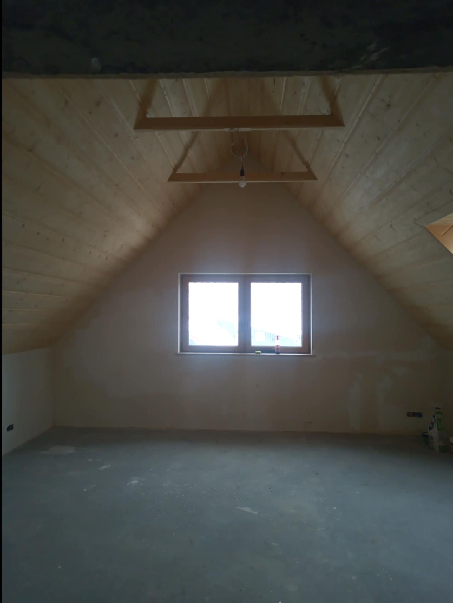 Poddasze z oknem po wstępnym wykończeniu: drewniana boazeria na skośnym suficie, gładkie ściany, surowa wylewka. Widoczne elementy instalacji i materiały budowlane.