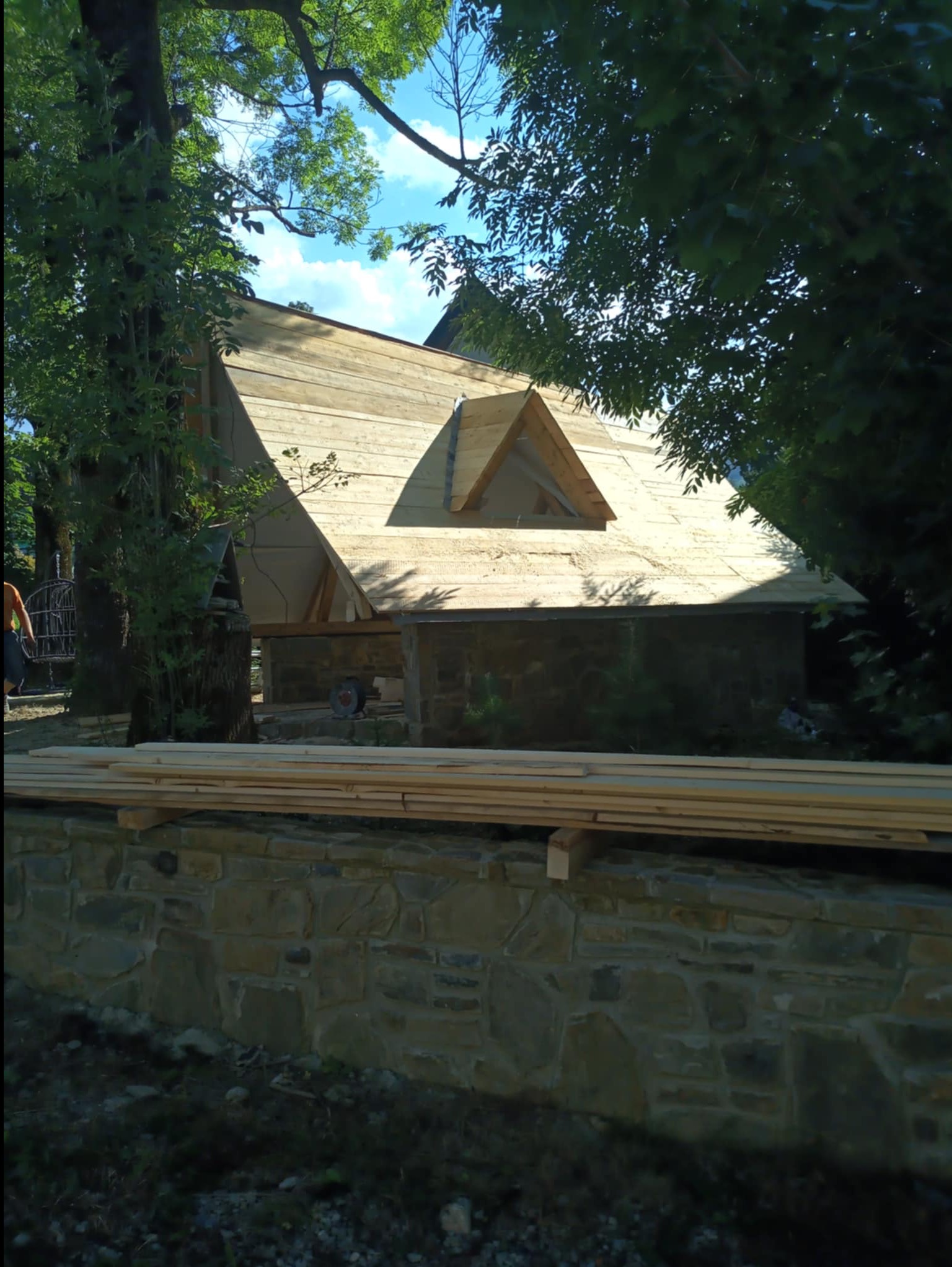 Budowa domu z bali w lesie: surowy dach z lukarną, kamienna podmurówka i stos desek na pierwszym planie. Widok z dołu, częściowo zasłonięty przez drzewa.