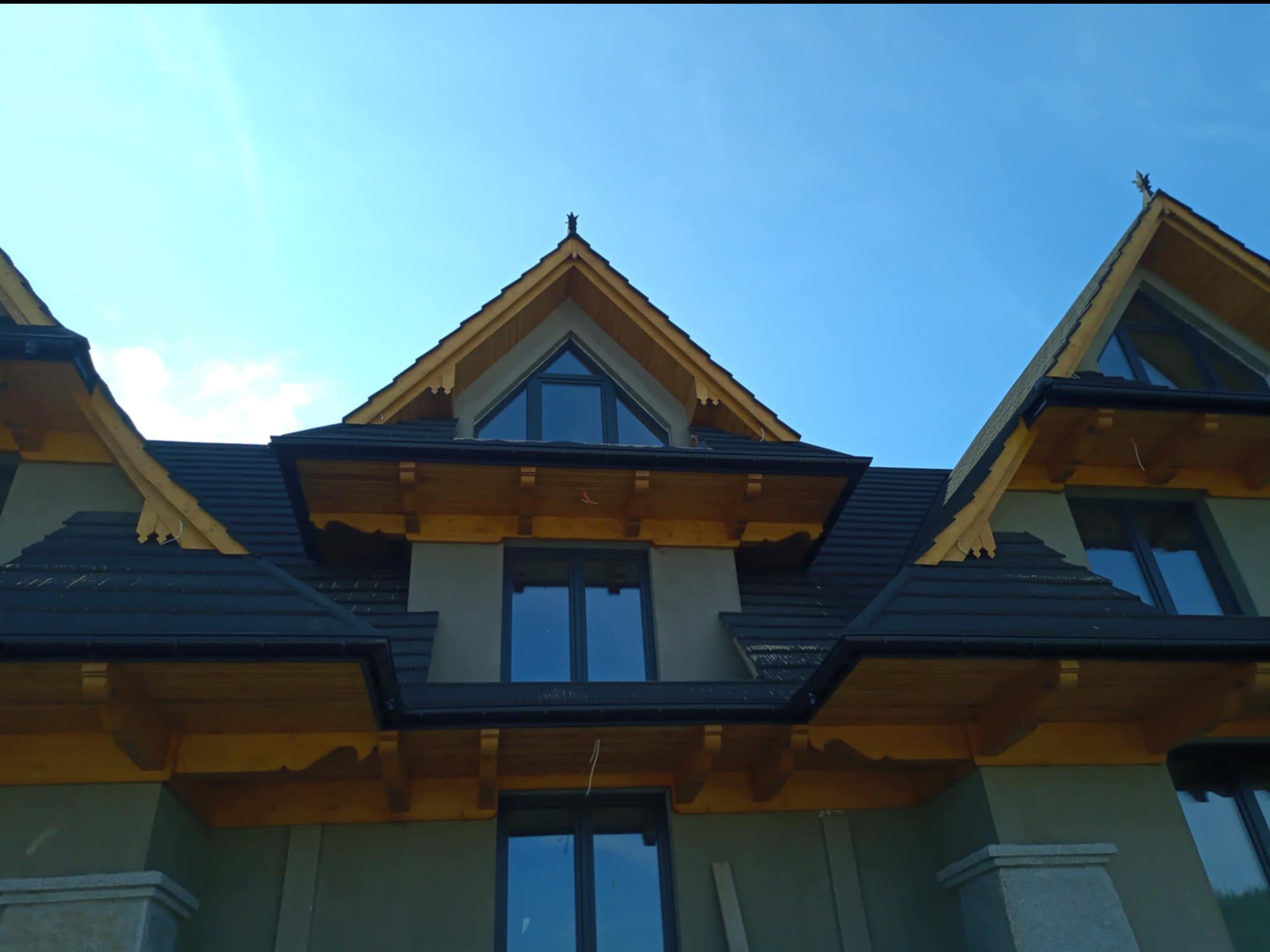 Fasada domu w stylu góralskim z ciemną dachówką i drewnianymi elementami wykończeniowymi, widziana z dołu pod kątem, na tle błękitnego nieba.