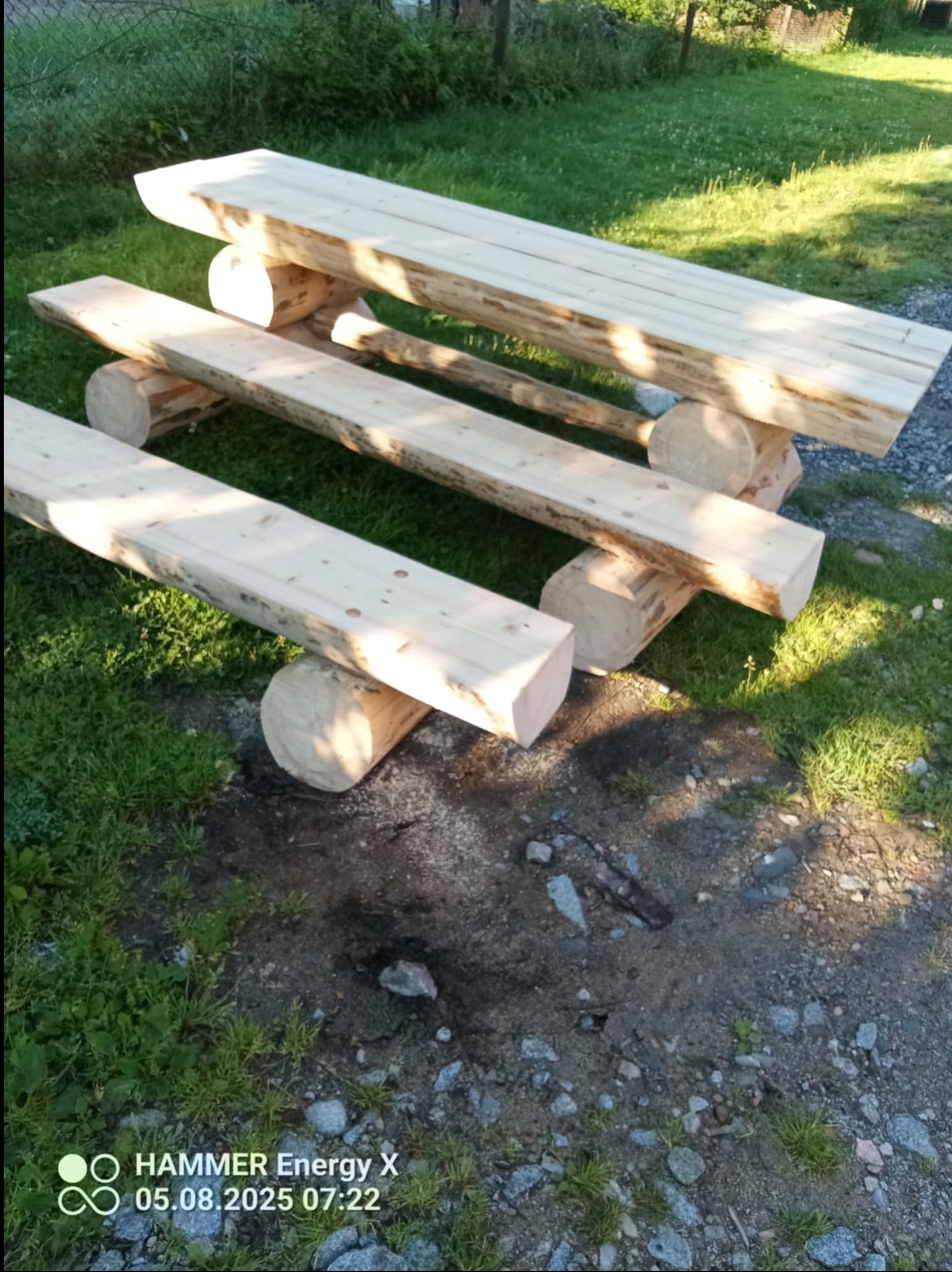 Surowy, drewniany stół i ławki z bali, ustawione na zewnątrz. Widoczne niedoskonałości drewna i naturalne otoczenie. Prosta konstrukcja, świeże drewno.