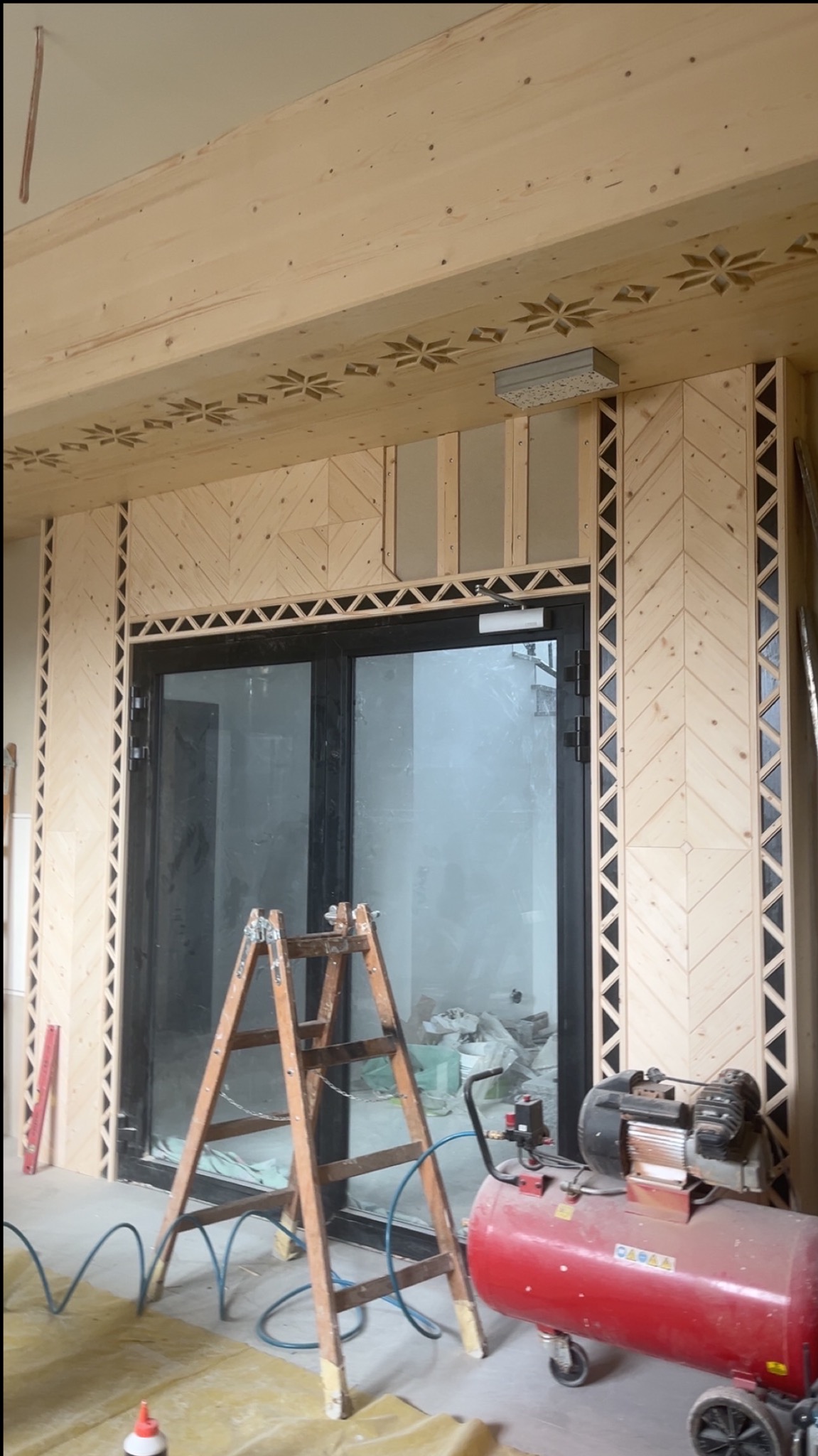 Drewniane wykończenie ściany z wzorem jodełki wokół czarnych drzwi, drabina i kompresor w pomieszczeniu w trakcie prac wykończeniowych.