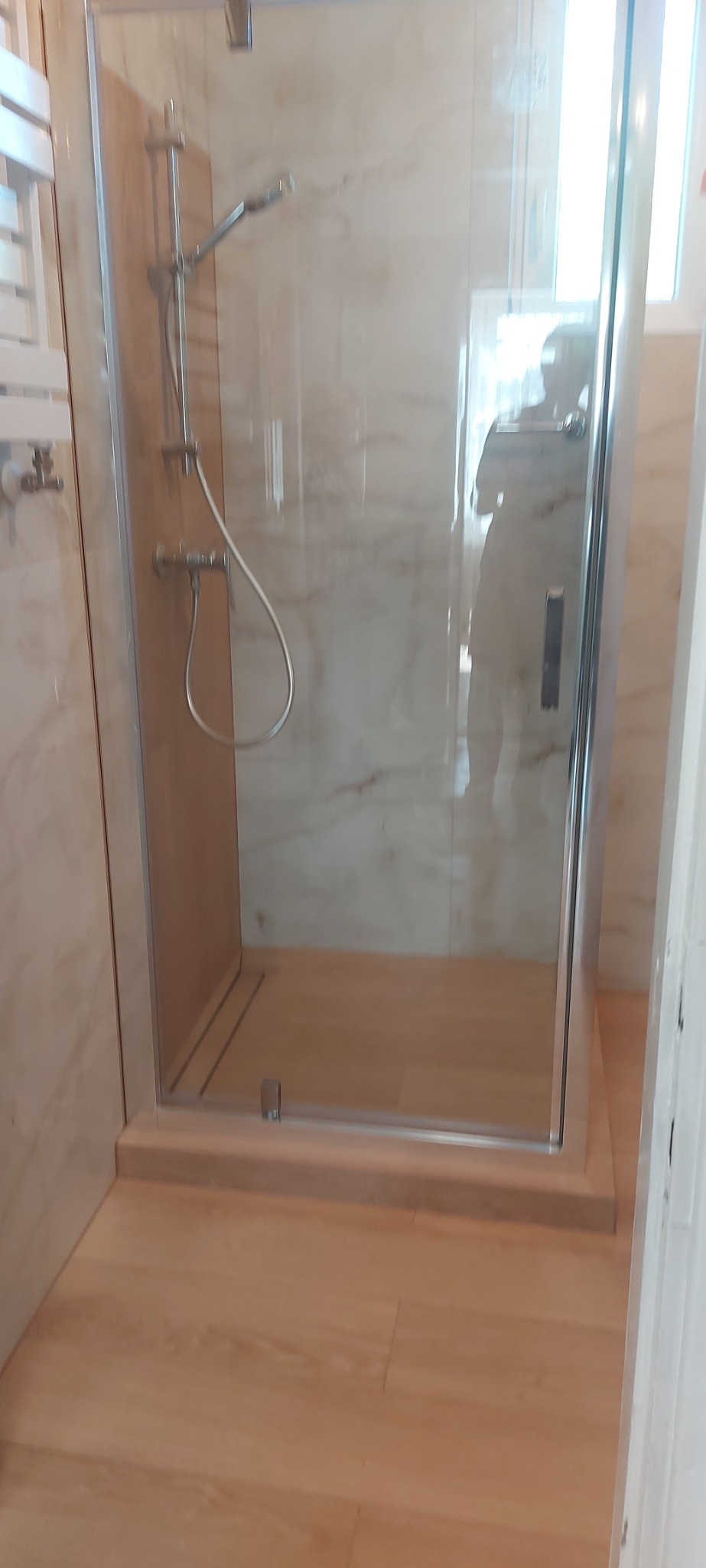 Szklana kabina prysznicowa z chromowaną armaturą na tle marmurowych ścian i drewnianej podłogi. Widoczne odbicie osoby w szybie.