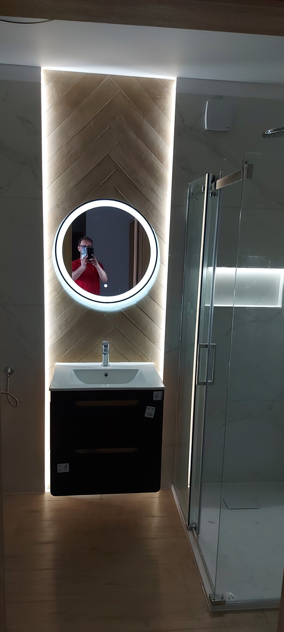 Nowoczesna łazienka z kabiną prysznicową i umywalką z szafką. Na ścianie lustro z podświetleniem LED i dekoracyjna okładzina w jodełkę. Widoczny fragment osoby robiącej zdjęcie.