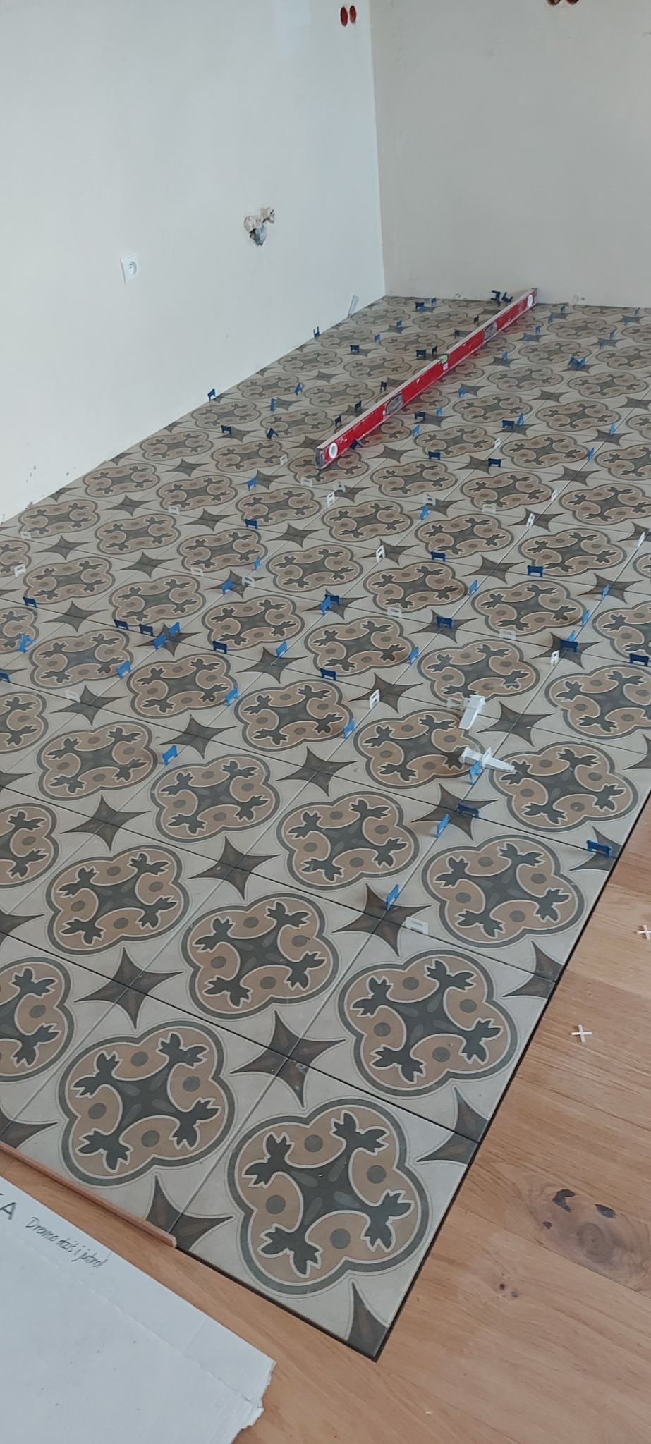 Układanie wzorzystych płytek podłogowych z użyciem poziomicy i klipsów dystansowych. Fragment drewnianej podłogi widoczny w rogu. Świeżo położona podłoga.