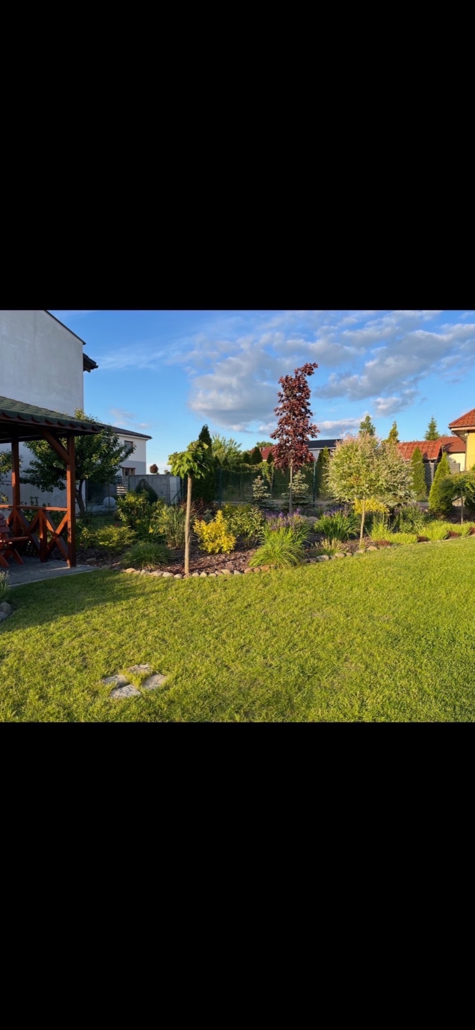 Zadbany ogród z równo przystrzyżonym trawnikiem, ozdobnymi drzewami i krzewami, altaną w rogu. Widok na domy w tle, błękitne niebo z chmurami.