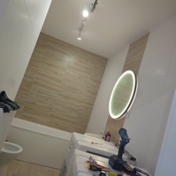 Wykończona łazienka z podświetlanym lustrem, biało-beżowymi płytkami i narzędziami na umywalce. Widoczne oświetlenie sufitowe i fragment wanny.