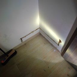 Przygotowanie pod instalację elektryczną w rogu pokoju z widocznymi kablami i bruzdą w ścianie, oświetloną punktowo. Na podłodze płytki imitujące drewno i szczotka.
