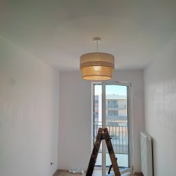 Wnętrze pokoju w trakcie wykańczania: drabina, lampa, grzejnik, wiertarka. Świeżo pomalowane ściany i widok z balkonu. Etap prac wykończeniowych.