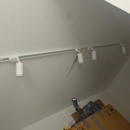 Biała szyna z reflektorami punktowymi zamontowana na skośnym suficie poddasza. Widoczne narzędzia na kartonie, sugerujące trwający montaż.
