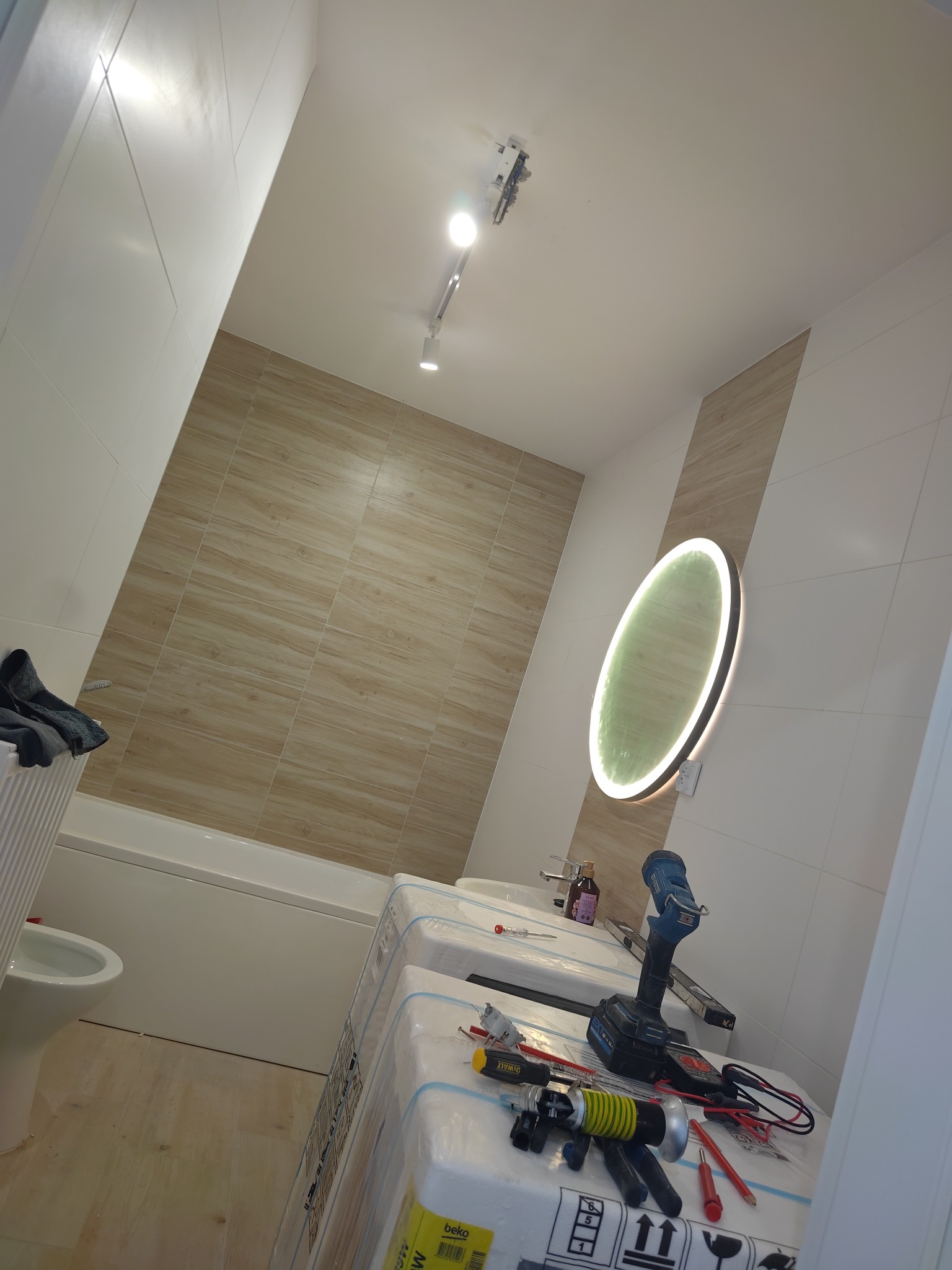 Wykończona łazienka z podświetlanym lustrem, biało-beżowymi płytkami i narzędziami na umywalce. Widoczne oświetlenie sufitowe i fragment wanny.