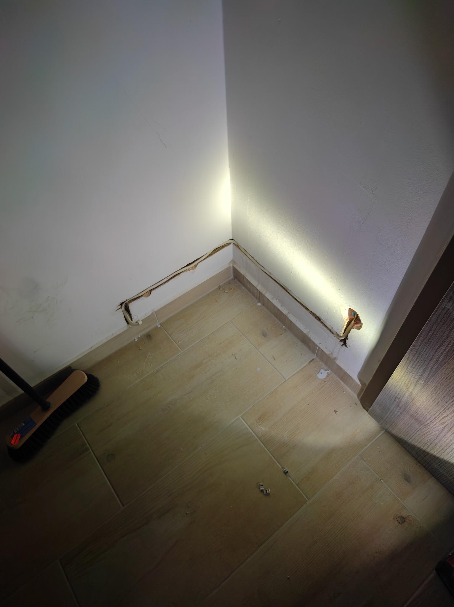 Przygotowanie pod instalację elektryczną w rogu pokoju z widocznymi kablami i bruzdą w ścianie, oświetloną punktowo. Na podłodze płytki imitujące drewno i szczotka.