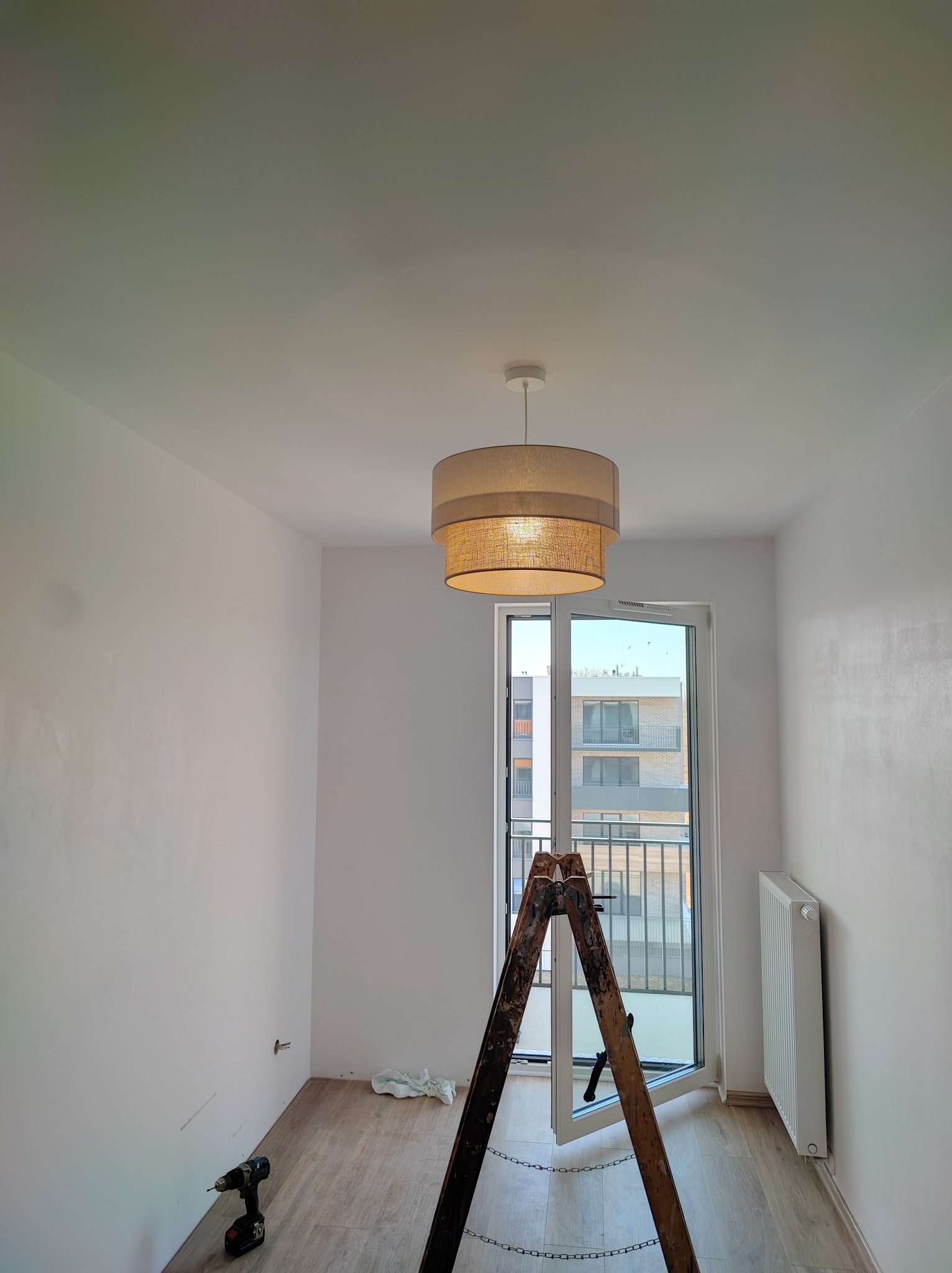 Wnętrze pokoju w trakcie wykańczania: drabina, lampa, grzejnik, wiertarka. Świeżo pomalowane ściany i widok z balkonu. Etap prac wykończeniowych.