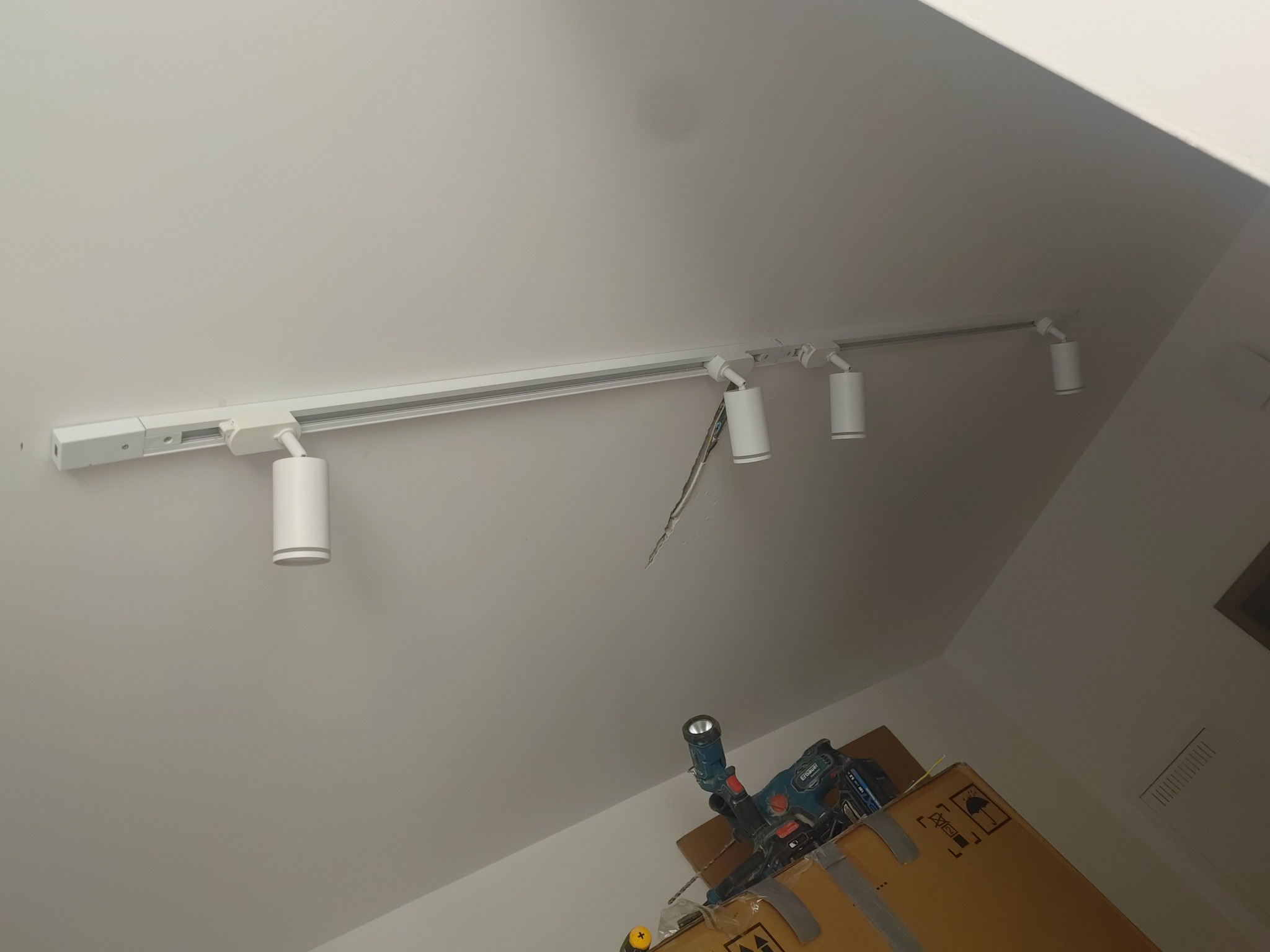 Biała szyna z reflektorami punktowymi zamontowana na skośnym suficie poddasza. Widoczne narzędzia na kartonie, sugerujące trwający montaż.