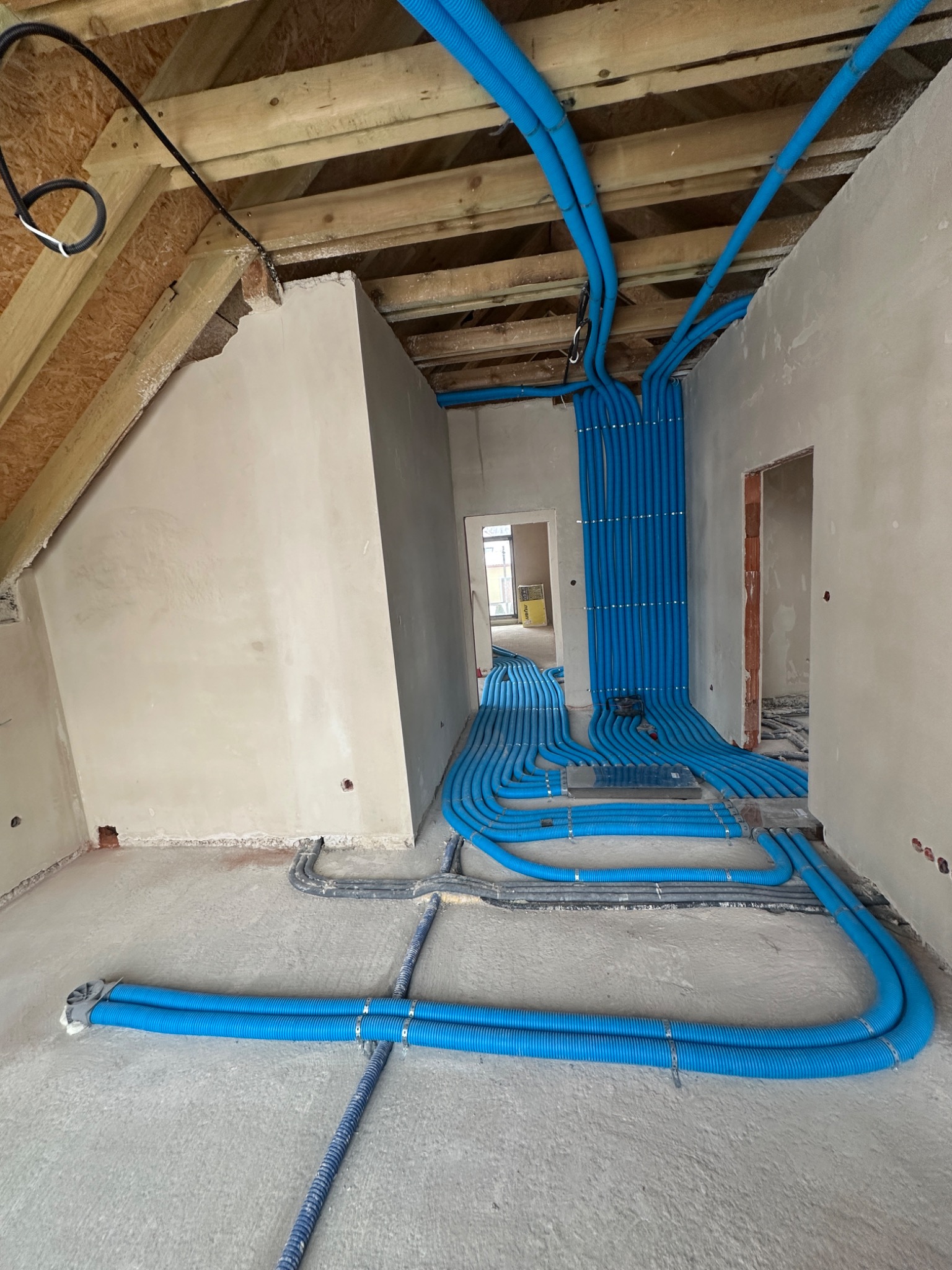 Niebieskie rury instalacji ogrzewania podłogowego ułożone w surowym wnętrzu poddasza, widoczne drewniane elementy konstrukcji dachu i ściany w trakcie wykończenia.