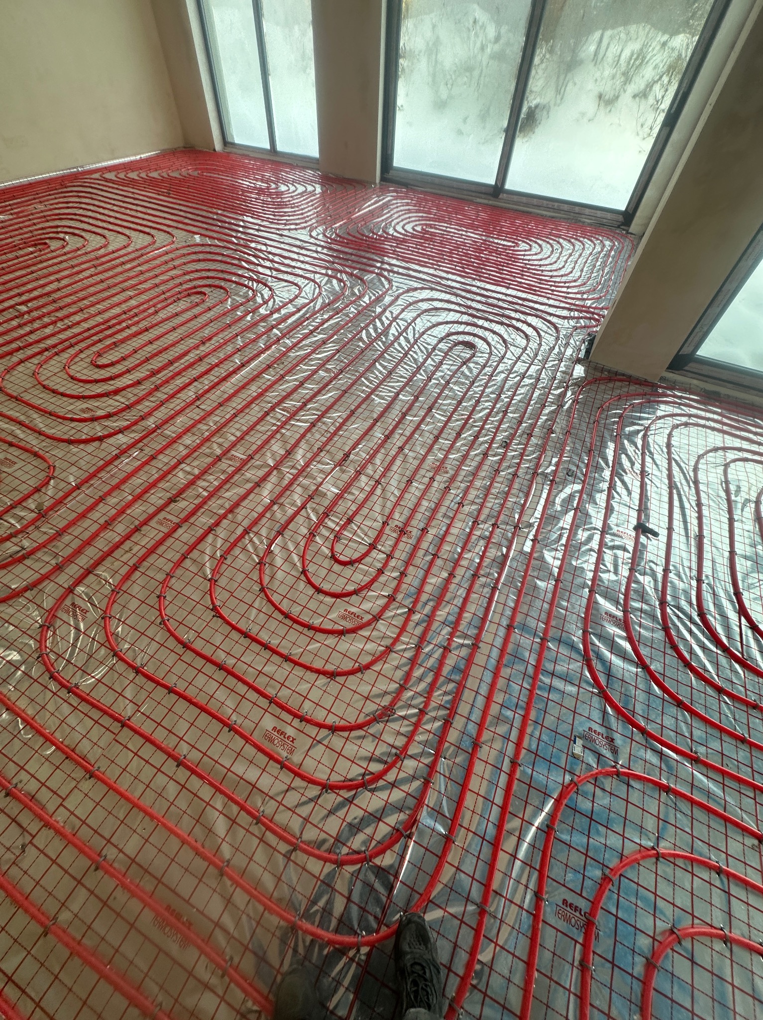 Czerwone rury ogrzewania podłogowego ułożone na folii refleksyjnej w jasnym pomieszczeniu z oknami. Widoczny fragment buta w rogu kadru.