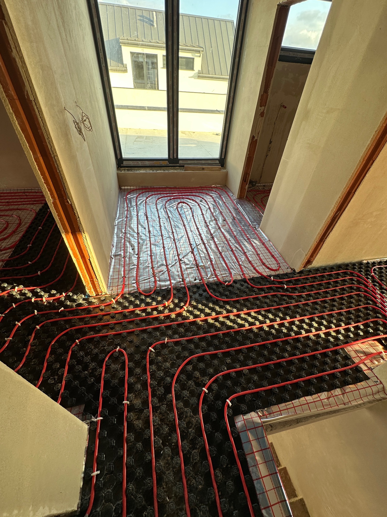Czerwone rury ogrzewania podłogowego ułożone na czarnej macie w jasnym pomieszczeniu z oknem dachowym, widok z góry, etap instalacji przed wylewką.