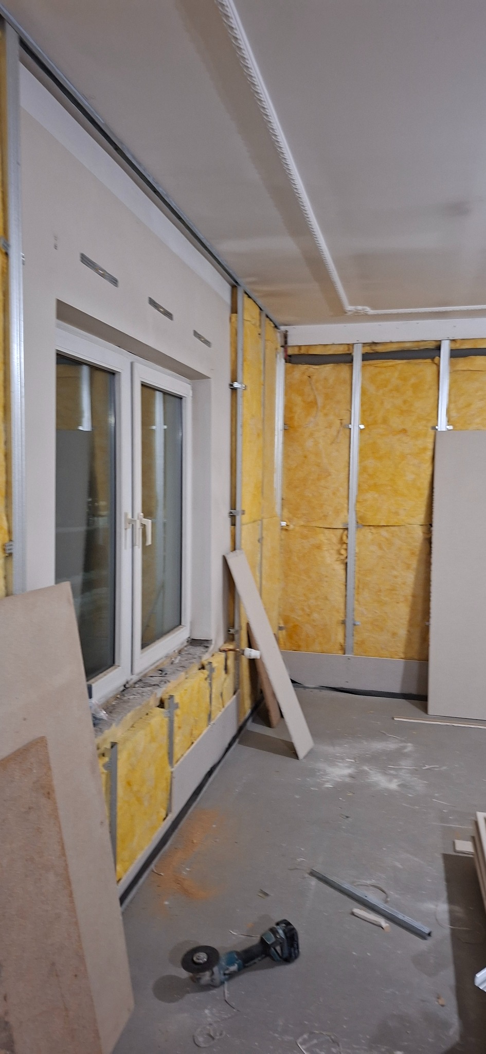 Wnętrze w trakcie prac wykończeniowych: izolacja ścian wełną mineralną, metalowy stelaż pod płyty gipsowe, widoczne narzędzia. Okno z białymi ramami.