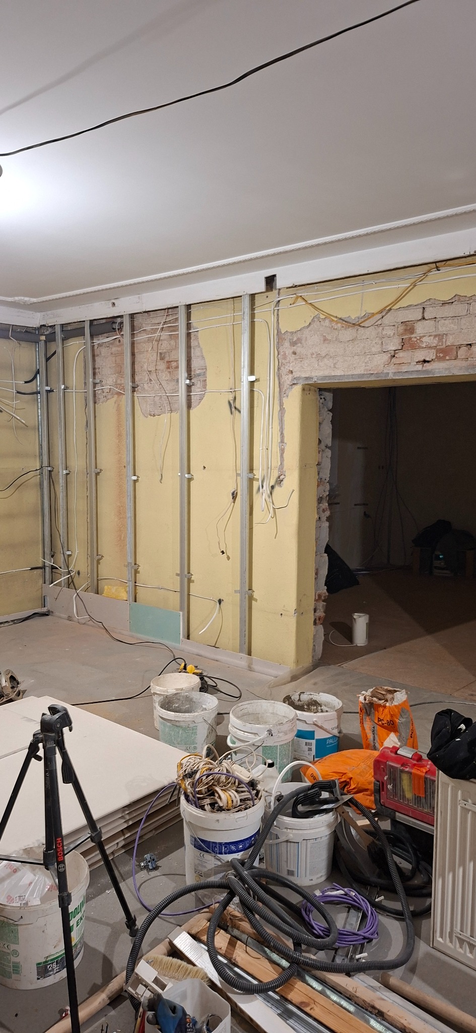 Wnętrze w trakcie remontu: metalowe profile na ścianie, widoczne instalacje elektryczne, surowe ściany, narzędzia i materiały budowlane. Etap przygotowania do prac wykończeniowych.